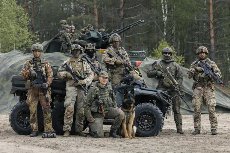 A group of military personnel in camouflage gear stands in front of a forest backdrop, posing with a K9 dog and an armored vehicle. The equipment includes rifles and a military ATV. The individuals have helmets and tactical gear, with trees and greenery in the background.