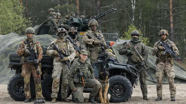 A group of military personnel in camouflage gear stands in front of a forest backdrop, posing with a K9 dog and an armored vehicle. The equipment includes rifles and a military ATV. The individuals have helmets and tactical gear, with trees and greenery in the background.
