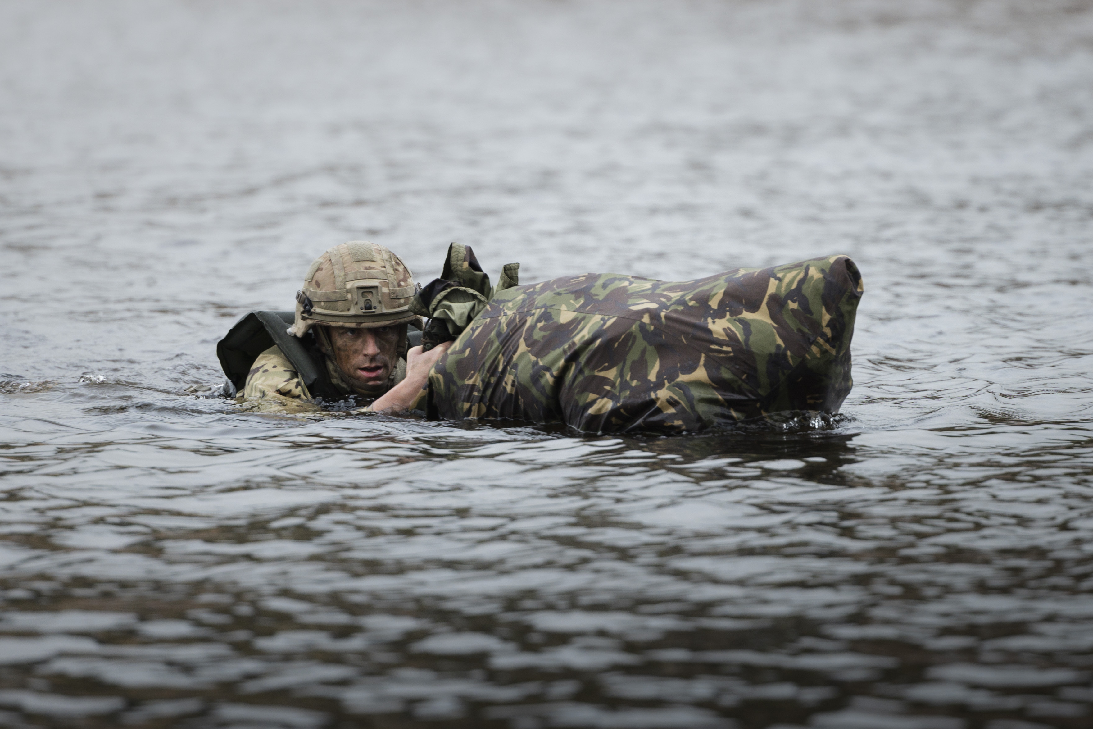 Soldier in camouflage swimming through water, holding a waterproof bag. 