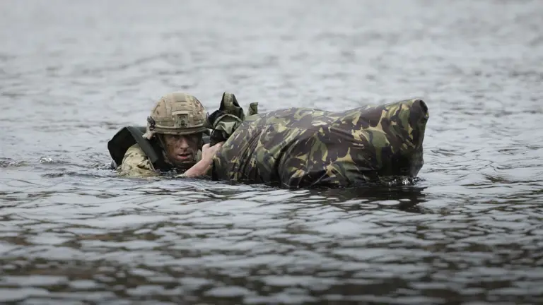 Soldier in camouflage swimming through water, holding a waterproof bag.