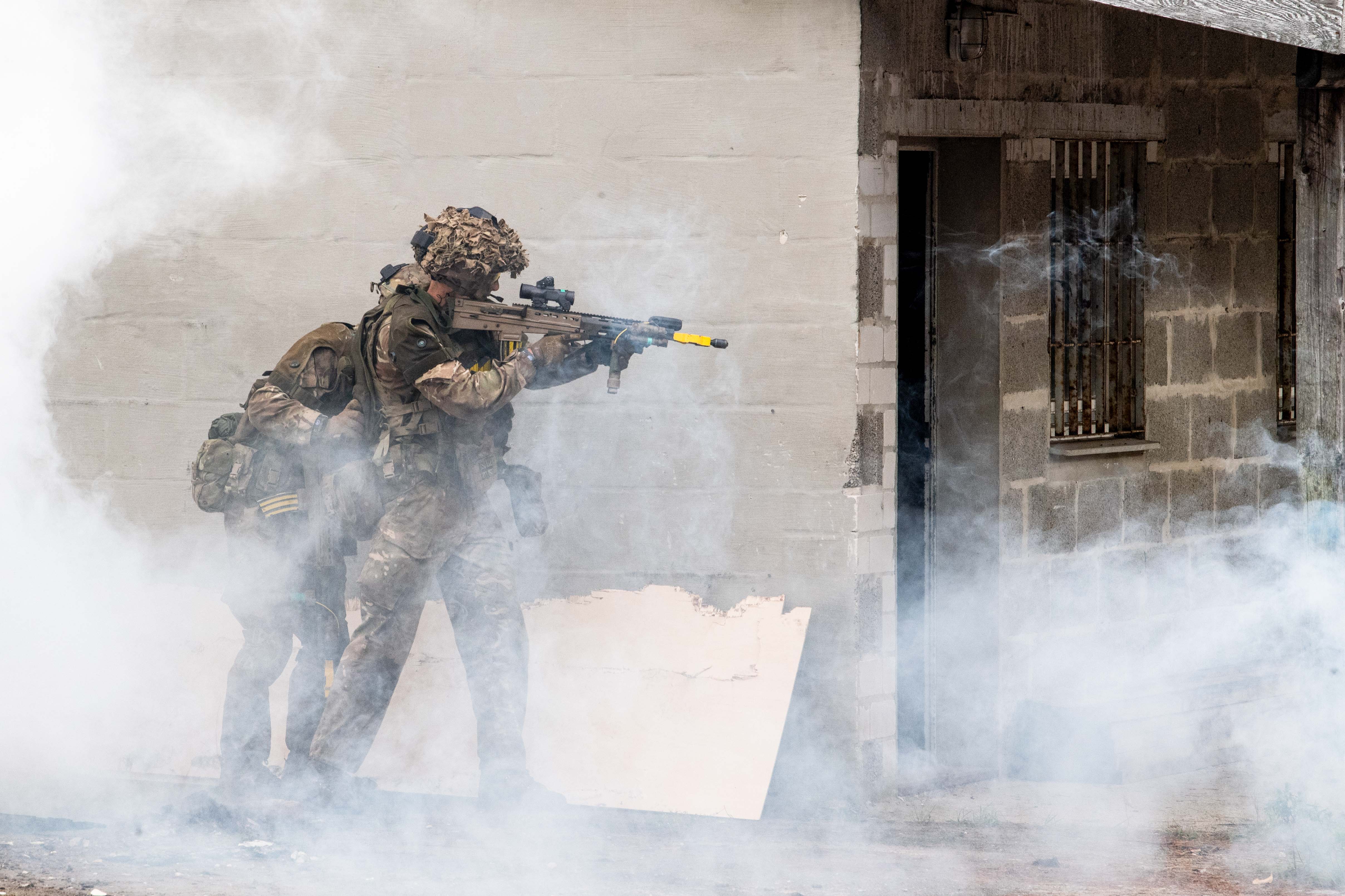 Two soldiers in tactical gear and helmets engage in a training exercise, moving beside a building. Thick smoke surrounds them.