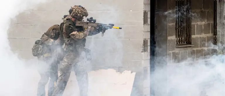 Two soldiers in tactical gear and helmets engage in a training exercise, moving beside a building. Thick smoke surrounds them.