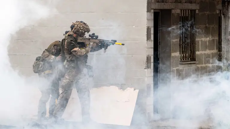 Two soldiers in tactical gear and helmets engage in a training exercise, moving beside a building. Thick smoke surrounds them.