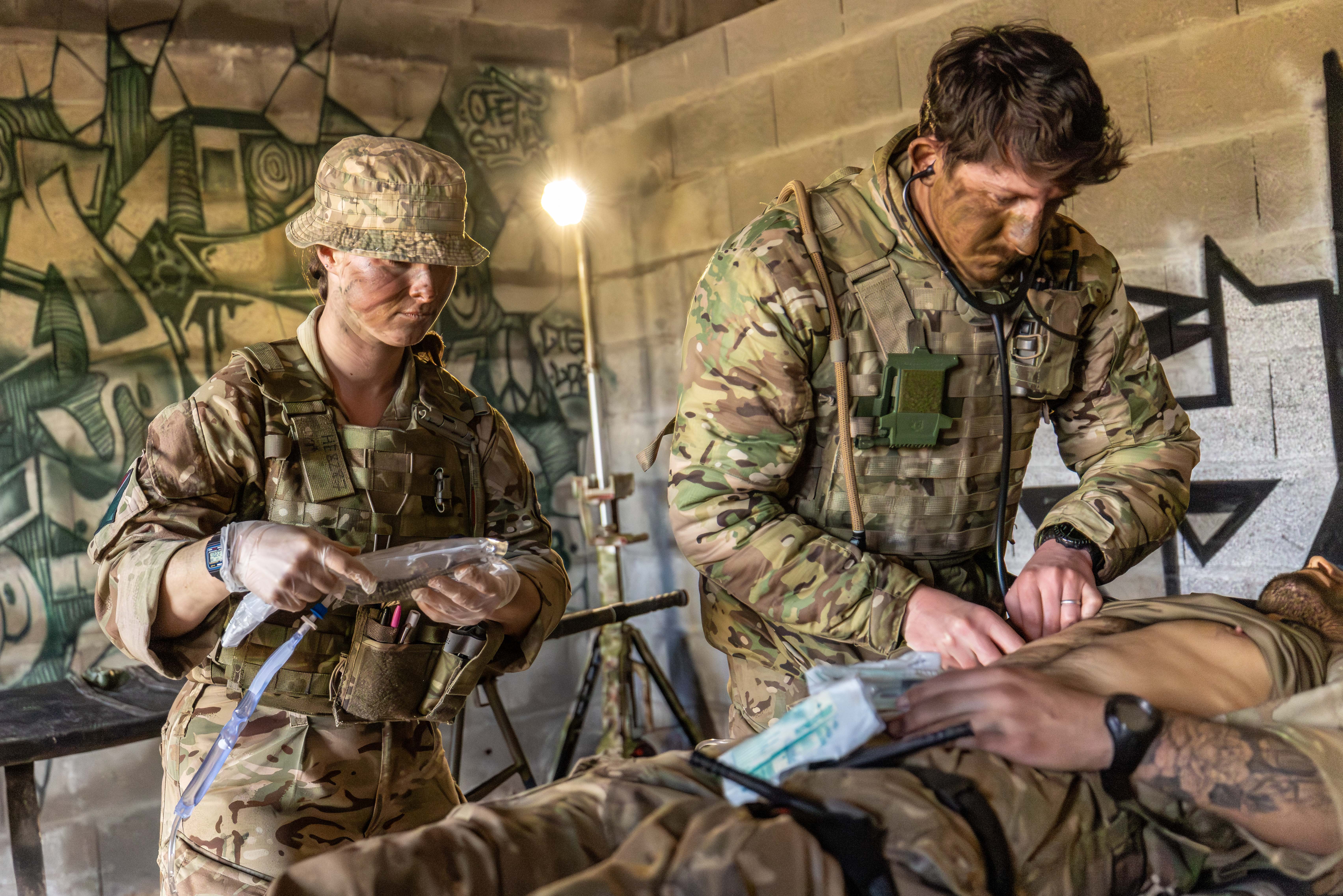 Two military personnel in camouflage uniforms perform a medical procedure in a room with graffiti on the walls. 