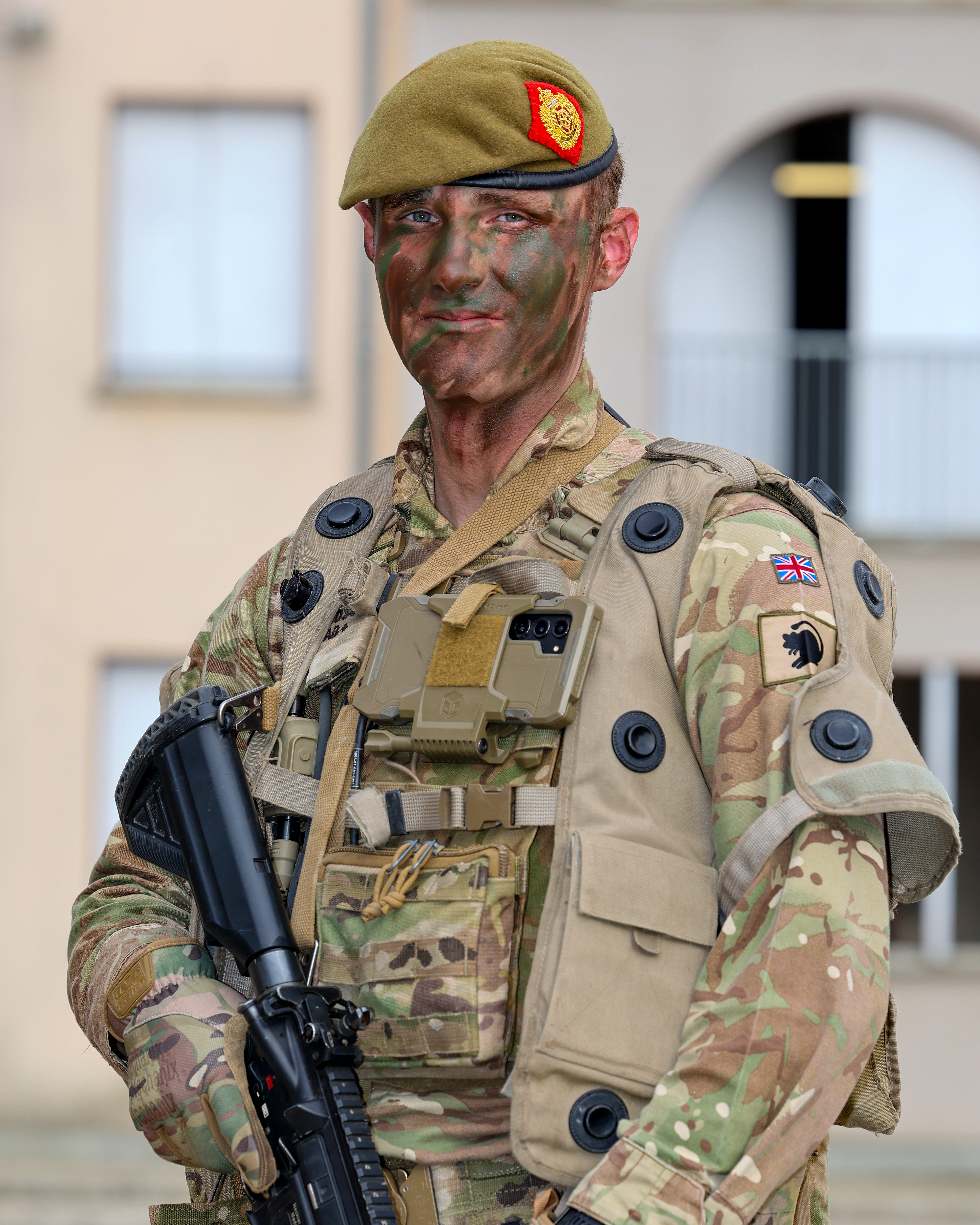 A person in camouflage military attire stands outdoors in front of a building. 