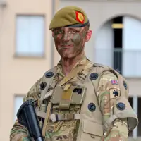 A person in camouflage military attire stands outdoors in front of a building.