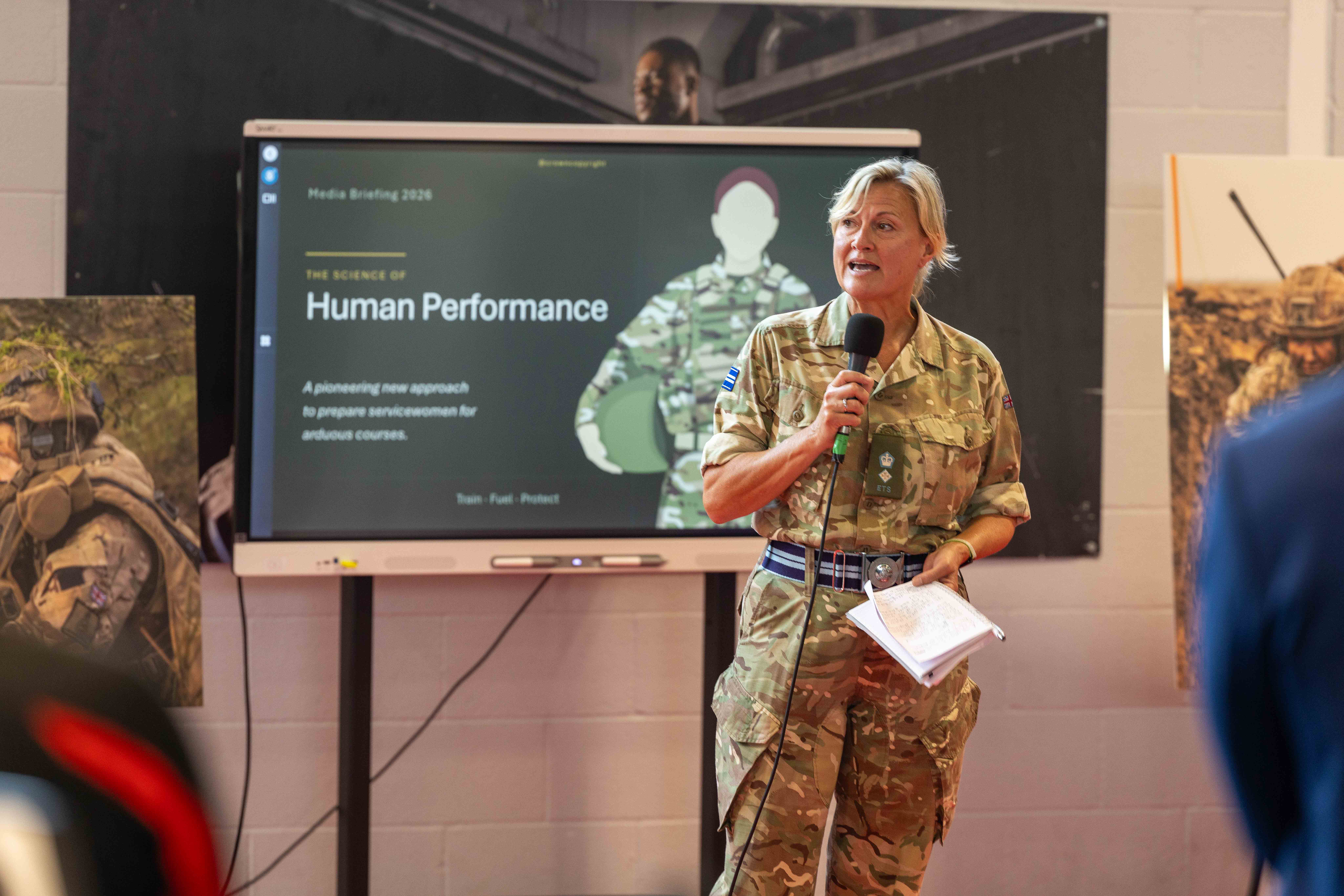 A person in military uniform stands holding a microphone in front of a presentation screen. 