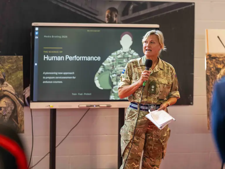 A person in military uniform stands holding a microphone in front of a presentation screen.