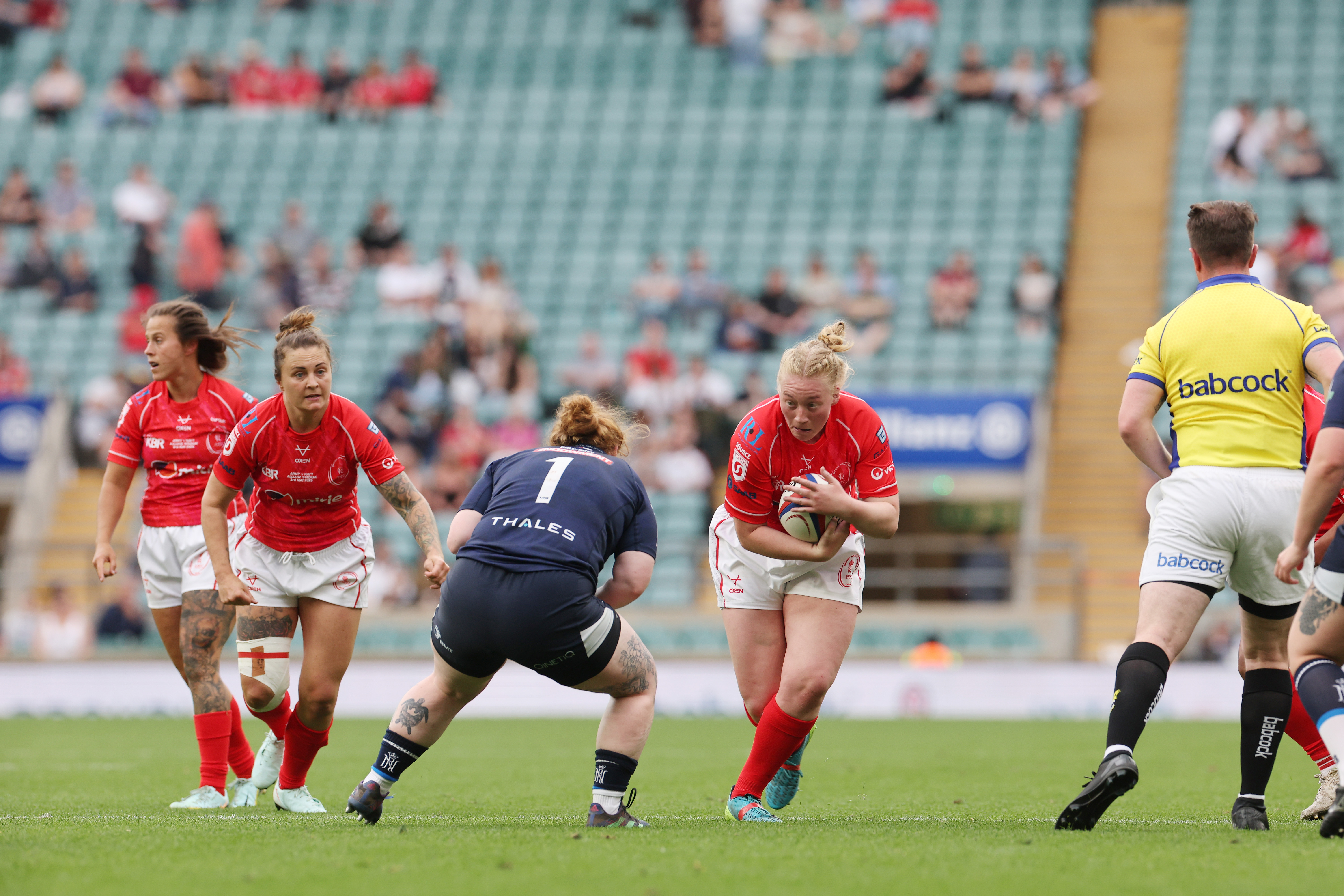 A women's rugby match in progress, player in red charges forward with the ball. Opponent in blue crouches to tackle. Teammates and referee nearby. Energetic atmosphere.