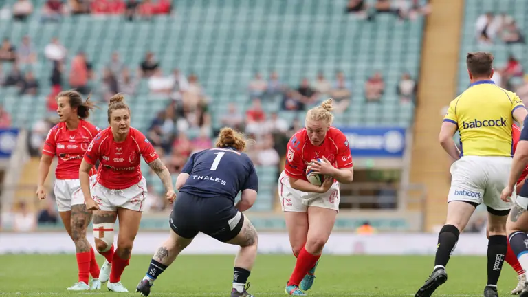 A women's rugby match in progress, player in red charges forward with the ball. Opponent in blue crouches to tackle. Teammates and referee nearby. Energetic atmosphere.
