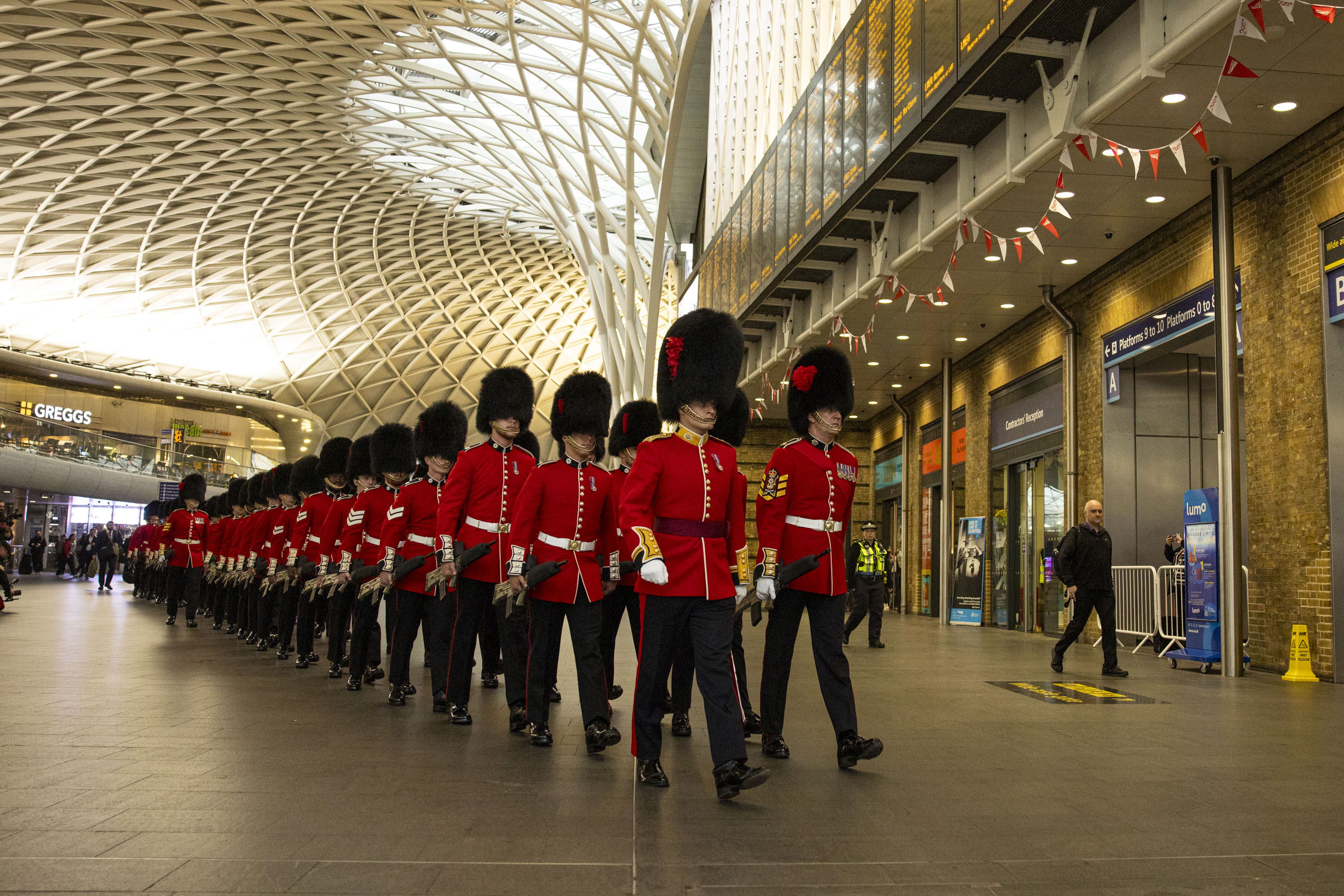 Soldiers wearing red tunics and black bearskin hats walk through Kings Cross Station.