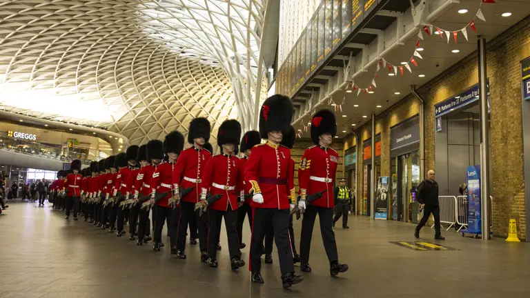 Soldiers wearing red tunics and black bearskin hats walk through Kings Cross Station.