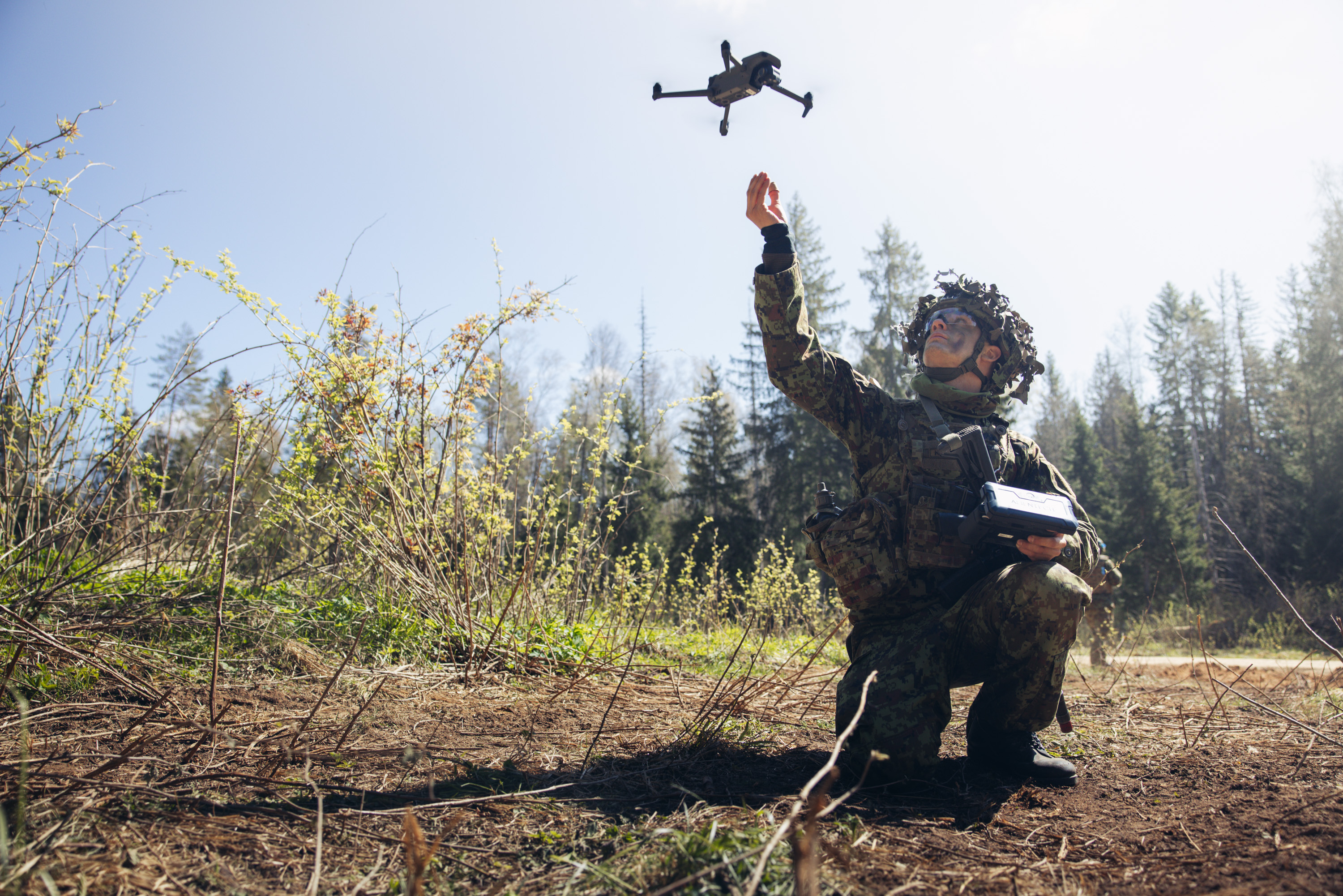 In a woodland clearing with trees in the background, a soldier is seen in camouflage uniform, kneeing while setting off a drone in the air with one hand and holding the controller in the other.