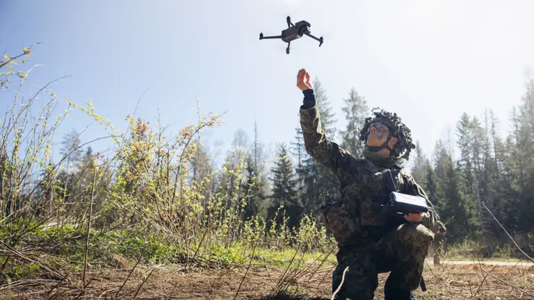 In a woodland clearing with trees in the background, a soldier is seen in camouflage uniform, kneeing while setting off a drone in the air with one hand and holding the controller in the other.