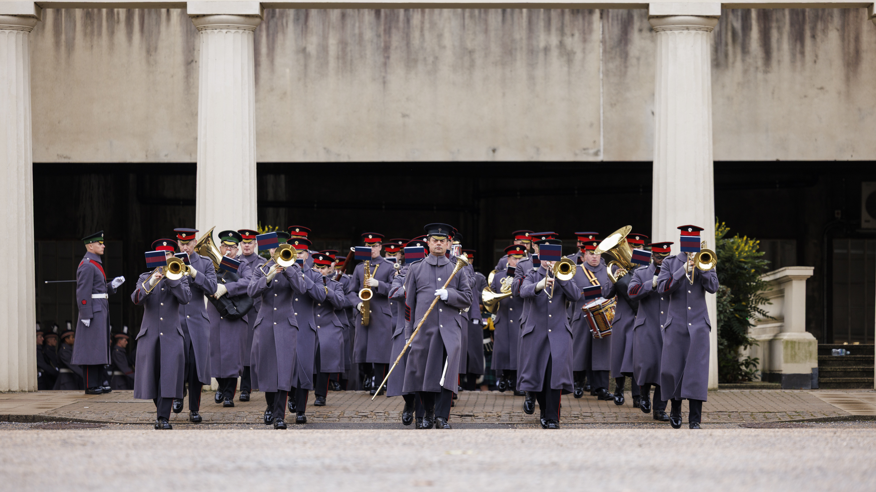 Military band in grey uniforms playing brass instruments whilst marching in formation outside a building with columns.