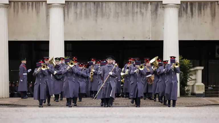 Military band in grey uniforms playing brass instruments whilst marching in formation outside a building with columns.