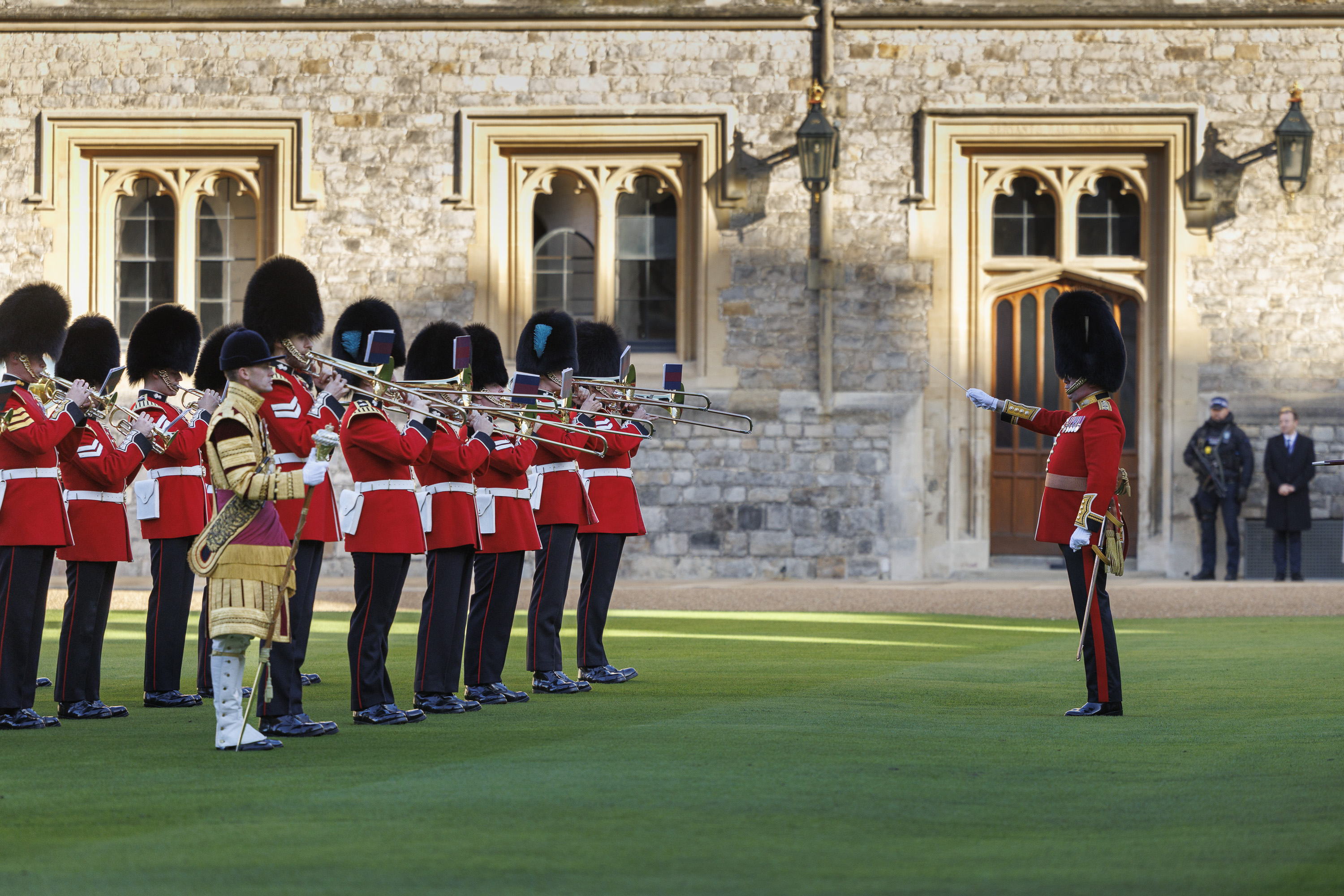 British royal guards in red uniforms and bearskin hats playing brass instruments during a ceremonial performance.