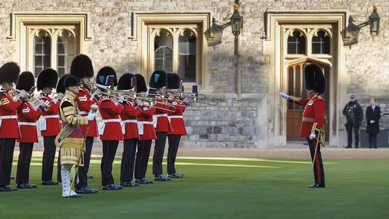 British royal guards in red uniforms and bearskin hats playing brass instruments during a ceremonial performance.