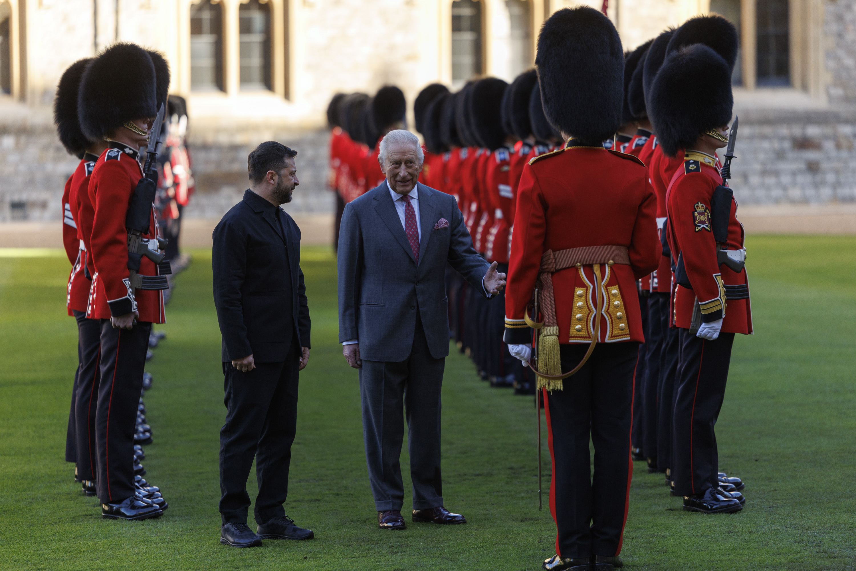 Two men stand on a lawn inspecting a line of British guards in traditional red uniforms and bearskin hats.