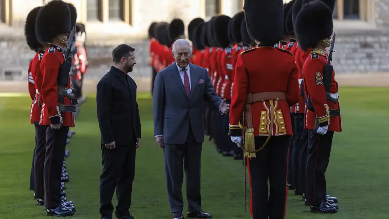 Two men stand on a lawn inspecting a line of British guards in traditional red uniforms and bearskin hats.