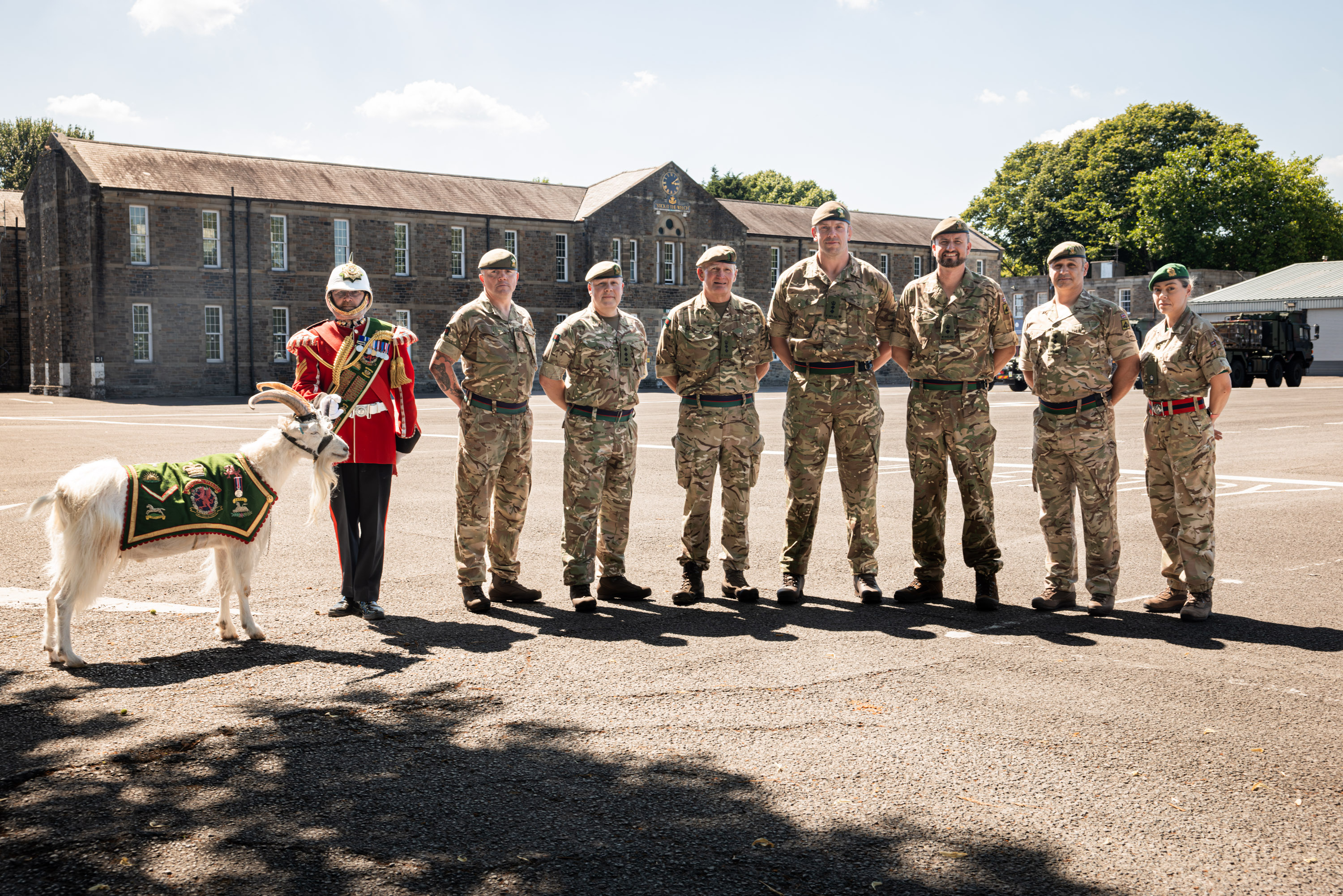 A row of eight people stand in front of a building. The person on the left is wearing a red ceremonial tunic and holding the lead of a goat. The rest of the people are wearing green camouflage uniforms.