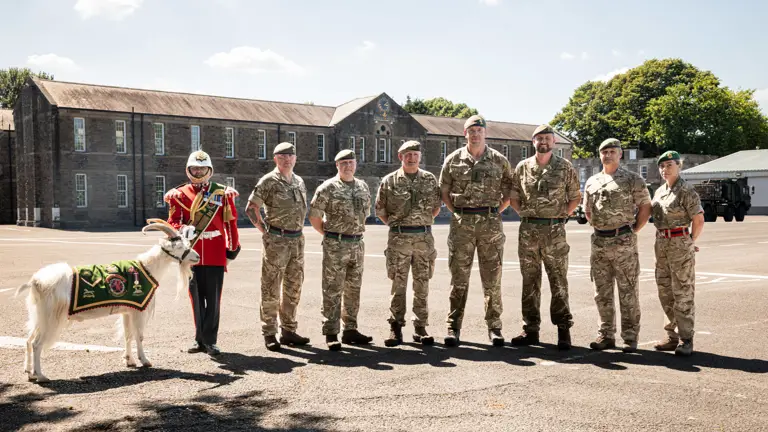 A row of eight people stand in front of a building. The person on the left is wearing a red ceremonial tunic and holding the lead of a goat. The rest of the people are wearing green camouflage uniforms.