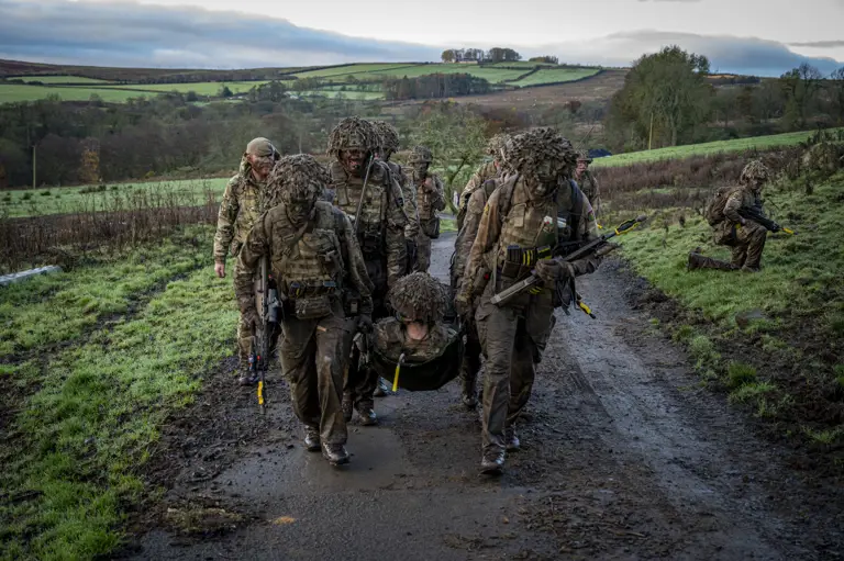 Soldiers wearing camouflage uniforms and carrying rifles carry a casualty up a hill.