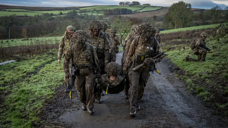 Soldiers wearing camouflage uniforms and carrying rifles carry a casualty up a hill.