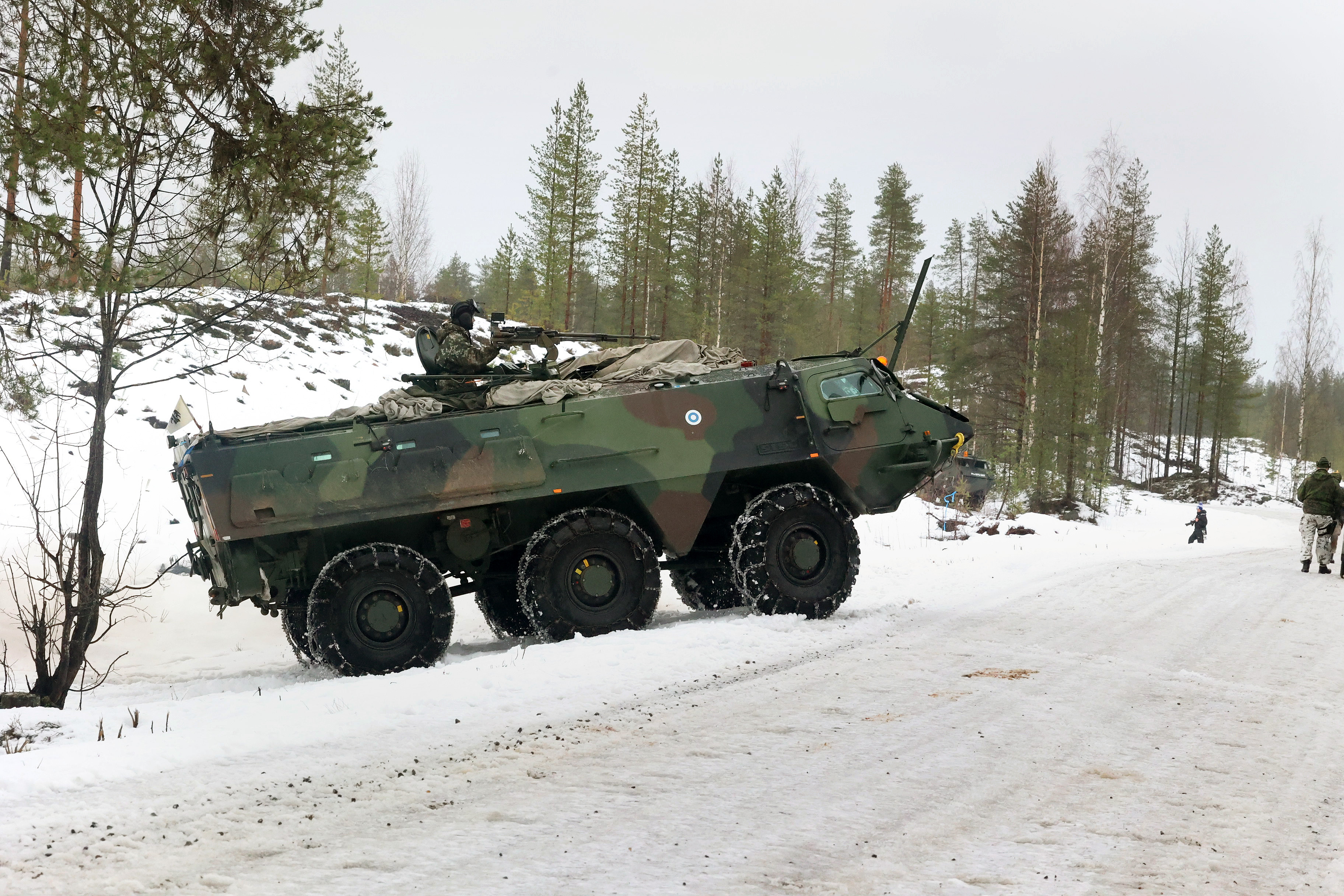 Camouflaged military vehicle on a snowy roadside, surrounded by tall evergreens. Soldiers in winter gear stand nearby, evoking a sense of readiness.