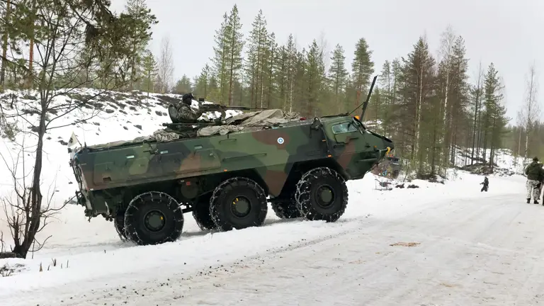 Camouflaged military vehicle on a snowy roadside, surrounded by tall evergreens. Soldiers in winter gear stand nearby, evoking a sense of readiness.