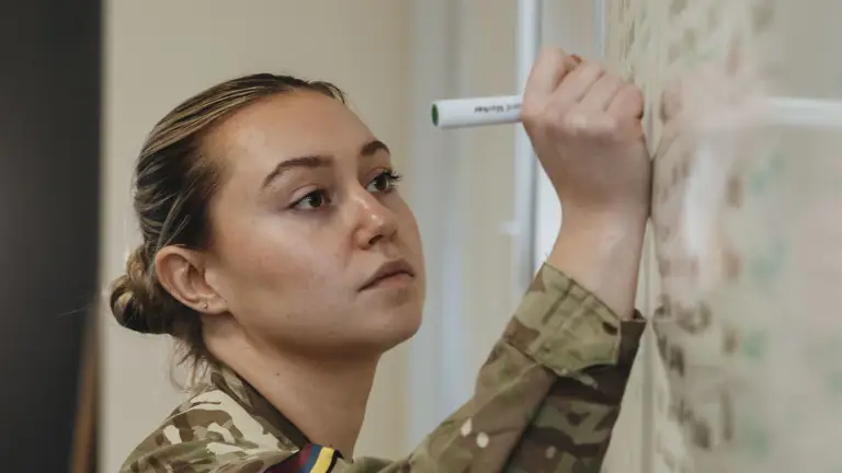 Military personnel in camouflage uniform writing notes on a whiteboard with a green marker in a classroom setting.