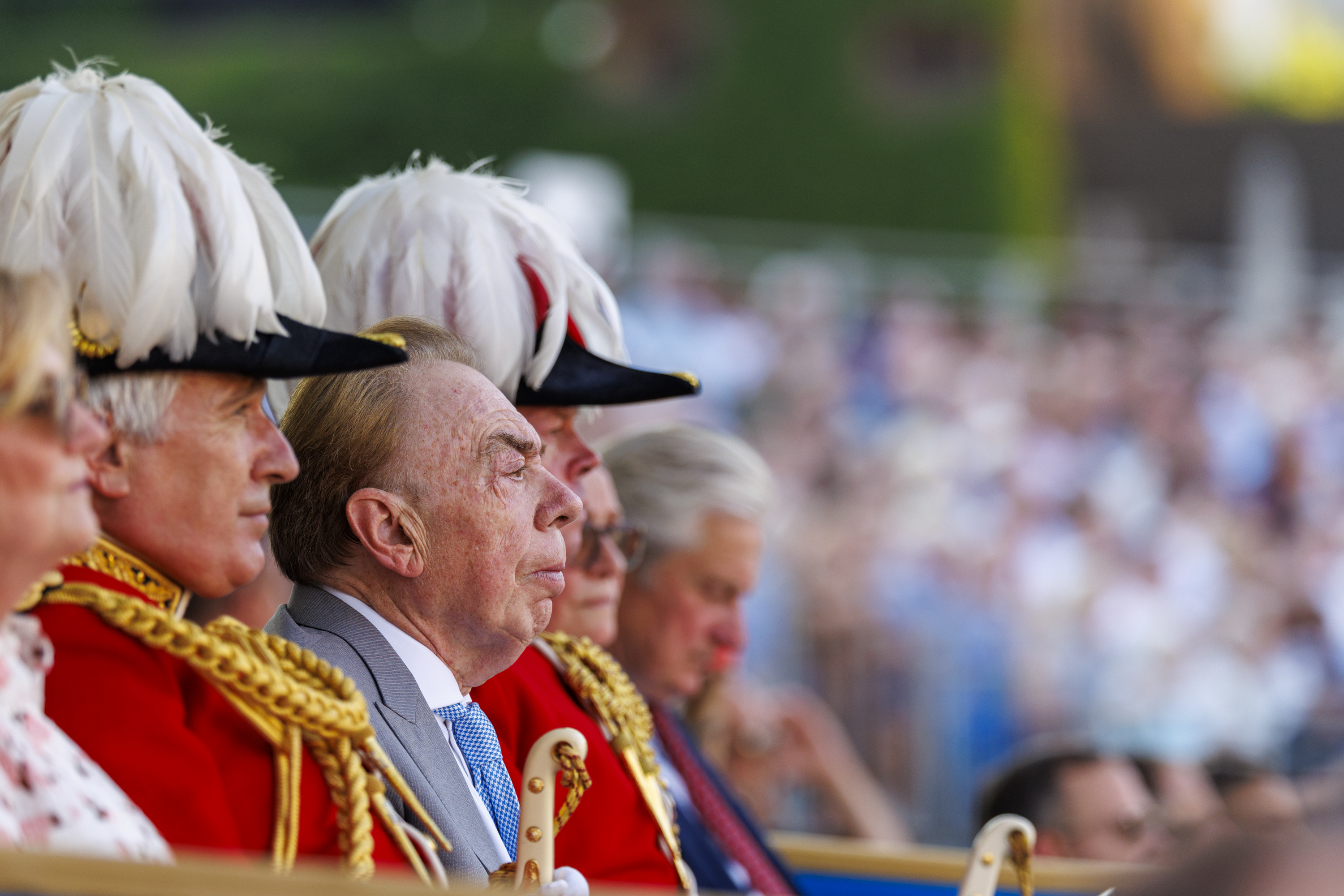 A group of people in formal red uniforms with gold detailing and white feathered hats sit together. One of them, possibly a public figure, is in focus. The background shows a blurred crowd, suggestive of a public event or ceremony.