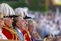 A group of people in formal red uniforms with gold detailing and white feathered hats sit together. One of them, possibly a public figure, is in focus. The background shows a blurred crowd, suggestive of a public event or ceremony.