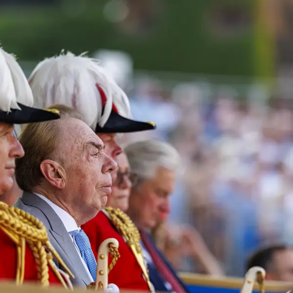 A group of people in formal red uniforms with gold detailing and white feathered hats sit together. One of them, possibly a public figure, is in focus. The background shows a blurred crowd, suggestive of a public event or ceremony.