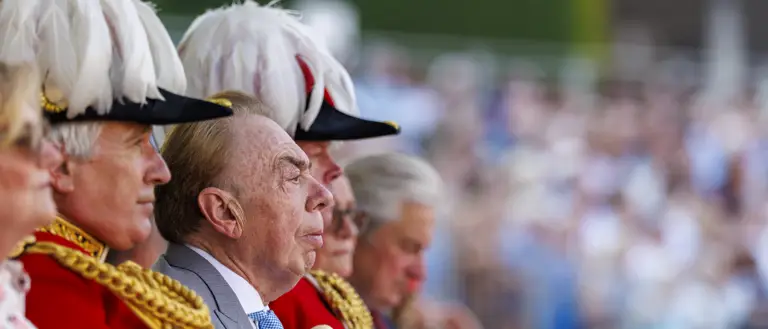 A group of people in formal red uniforms with gold detailing and white feathered hats sit together. One of them, possibly a public figure, is in focus. The background shows a blurred crowd, suggestive of a public event or ceremony.