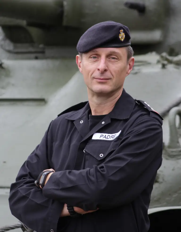 Reverend Mark Nightingale, wearing his black RTR coveralls and beret is stood posing arms folded in front of a tank.