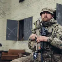 A soldier in uniform is seated outside of a building with his weapon.