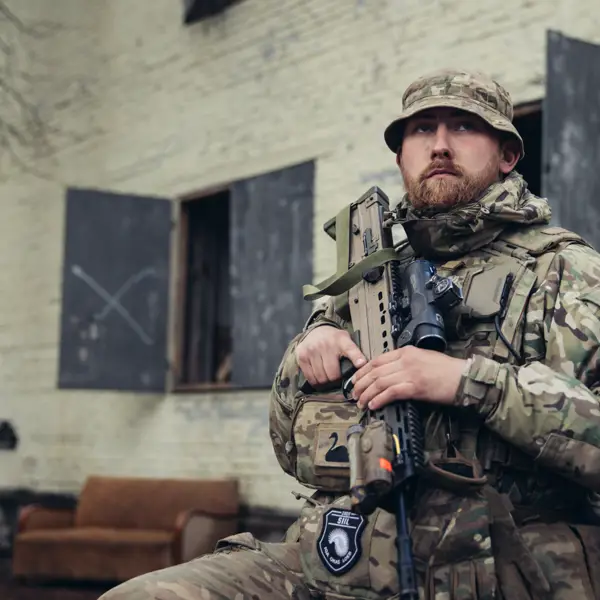 A soldier in uniform is seated outside of a building with his weapon.