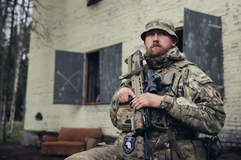 A soldier in uniform is seated outside of a building with his weapon.
