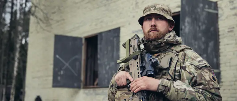 A soldier in uniform is seated outside of a building with his weapon.