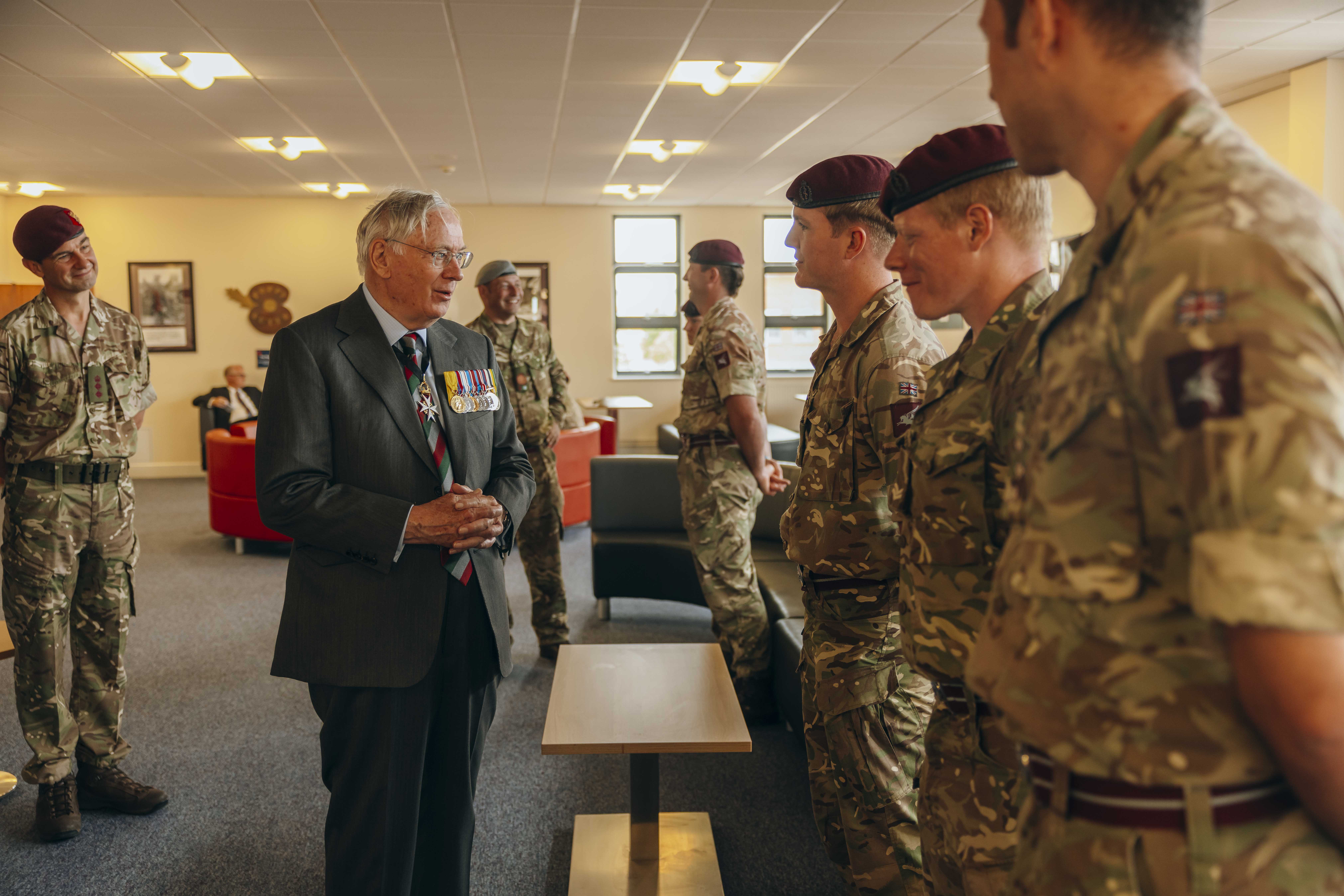 His Royal Highness the Duke of Gloucester stands wearing a suit. He is talking to three soldiers all wearing camouflage uniforms and maroon berets.