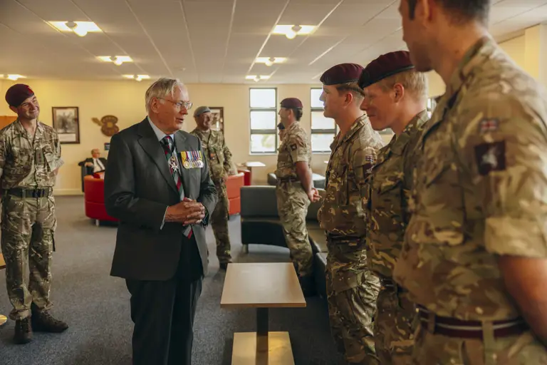 His Royal Highness the Duke of Gloucester stands wearing a suit. He is talking to three soldiers all wearing camouflage uniforms and maroon berets.