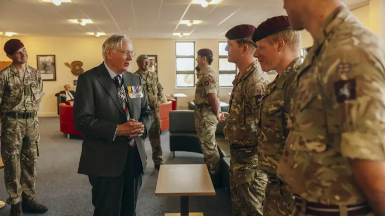 His Royal Highness the Duke of Gloucester stands wearing a suit. He is talking to three soldiers all wearing camouflage uniforms and maroon berets.