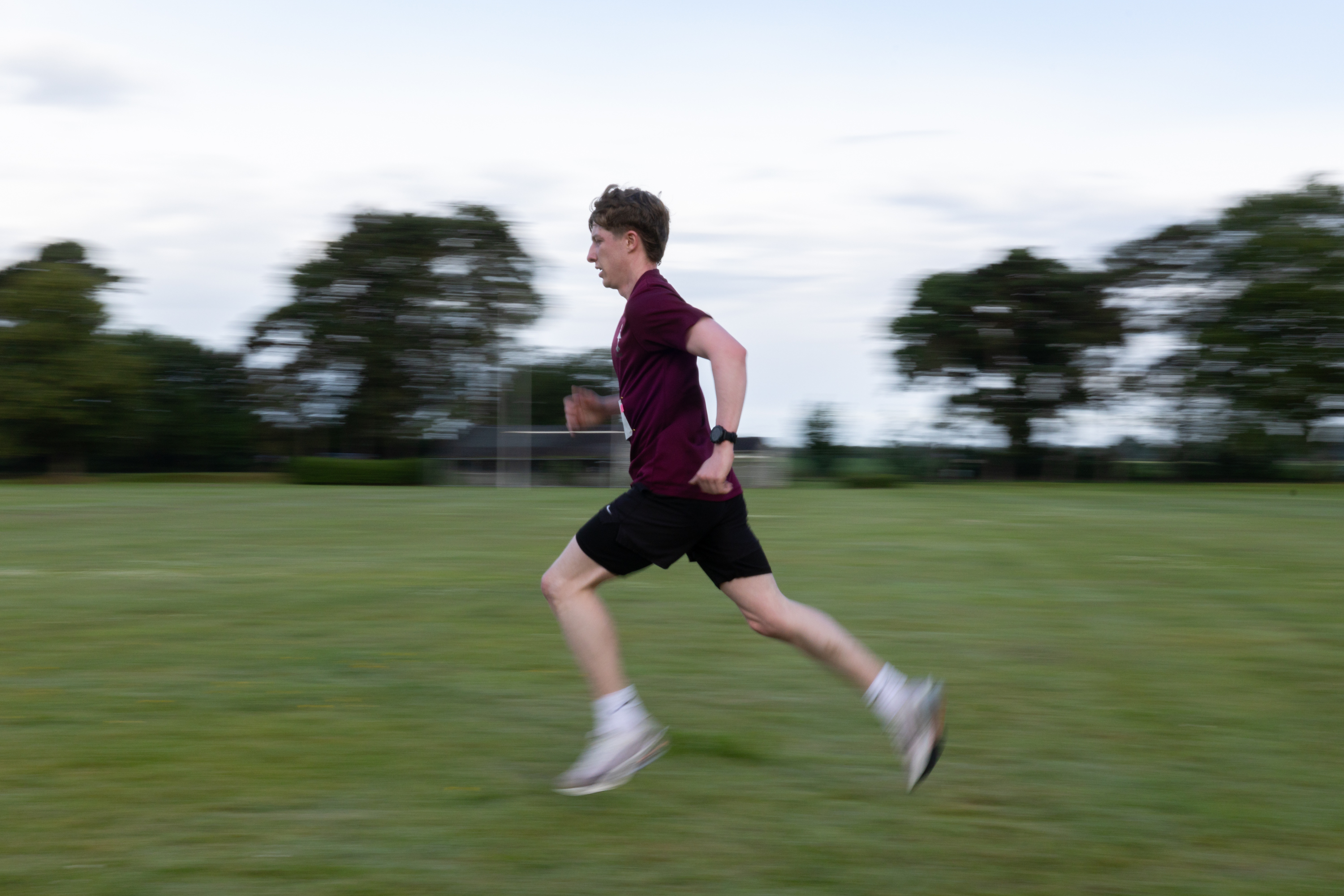 A person is running across a grassy field. Trees and a partially cloudy sky are in the background.