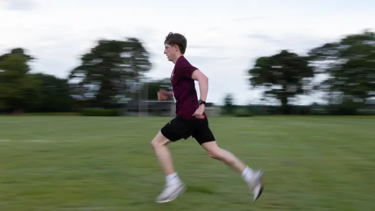A person is running across a grassy field. Trees and a partially cloudy sky are in the background.