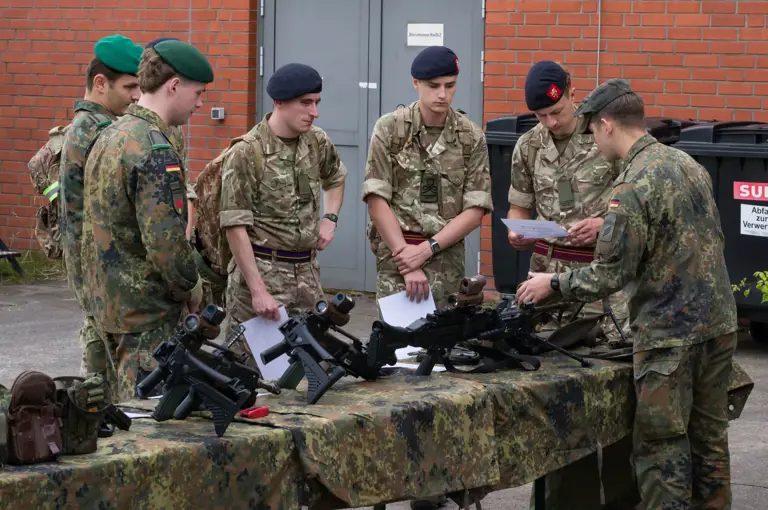 A group of people in military uniforms are gathered around a camouflaged table displaying various firearms. They appear to be in discussion while examining documents. A red brick wall is in the background.