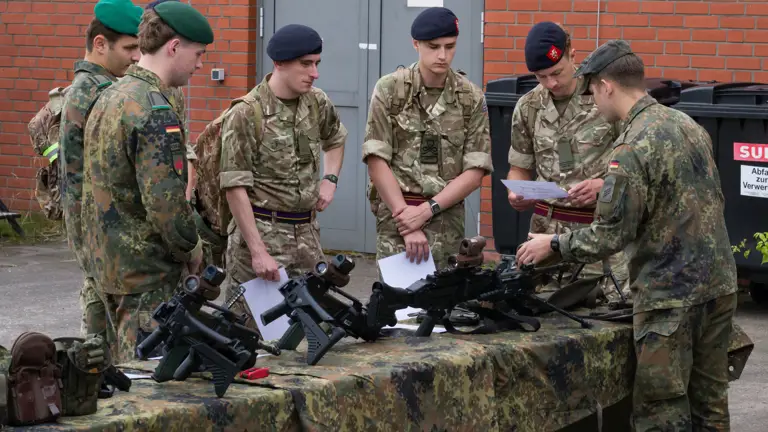 A group of people in military uniforms are gathered around a camouflaged table displaying various firearms. They appear to be in discussion while examining documents. A red brick wall is in the background.