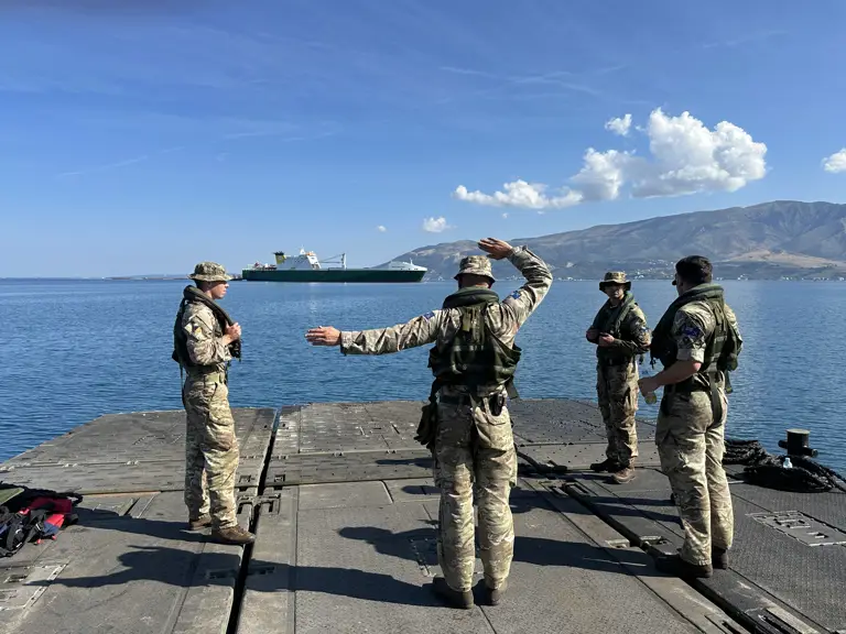 Four soldiers stand on a green military ferry whilst directing a ferry on the horizon.