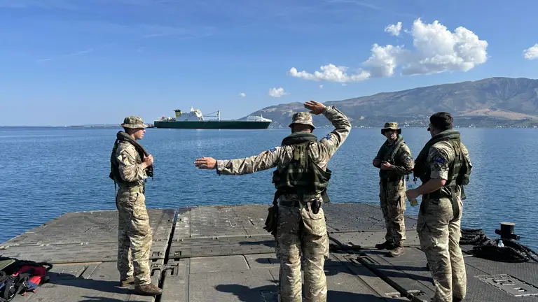 Four soldiers stand on a green military ferry whilst directing a ferry on the horizon.