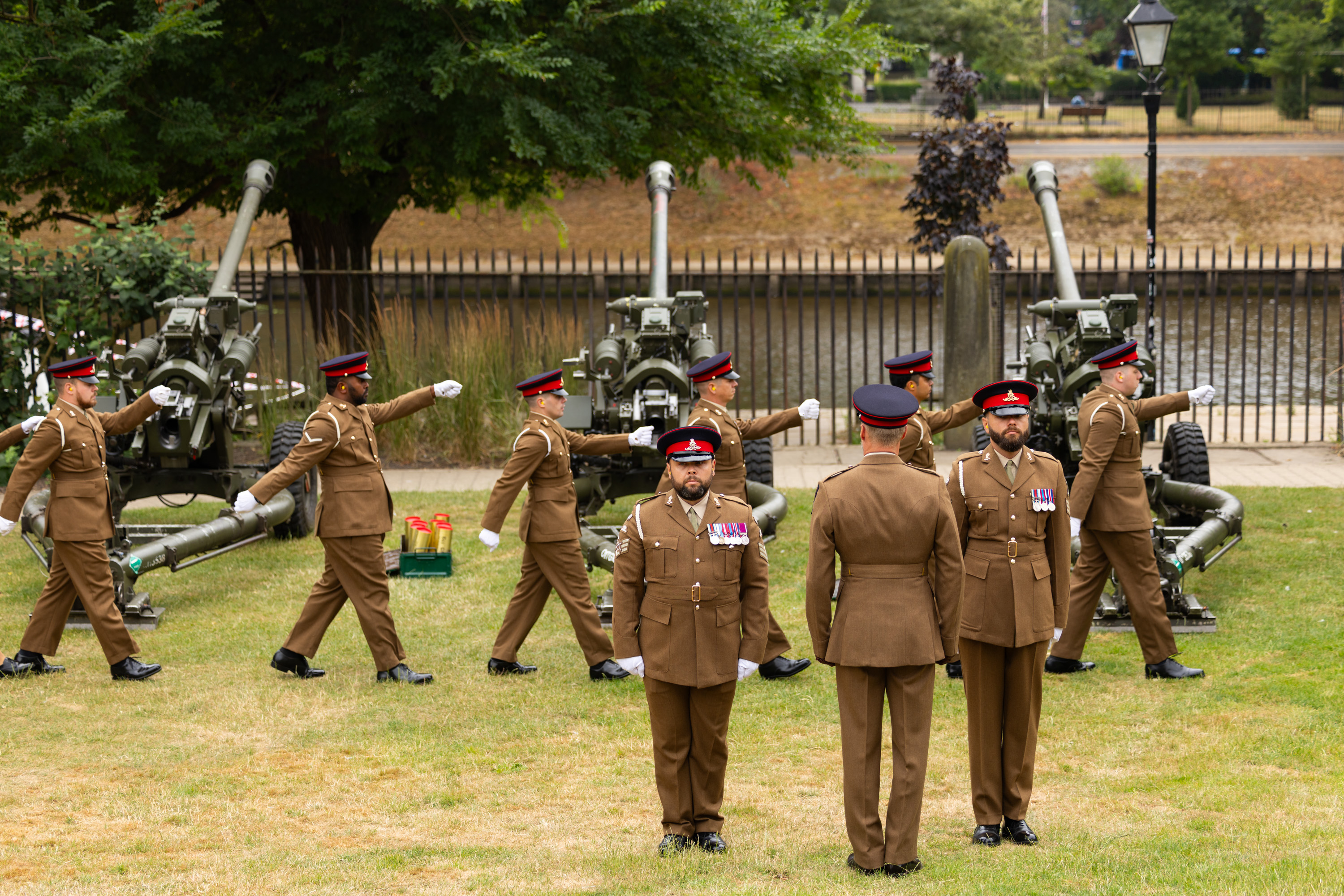 A group of uniformed soldiers stands and walks in formation on a grassy area in front of ceremonial cannons. 